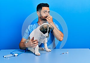 Handsome hispanic veterinary man with beard checking dog health smelling something stinky and disgusting, intolerable smell,