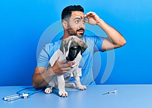 Handsome hispanic veterinary man with beard checking dog health very happy and smiling looking far away with hand over head