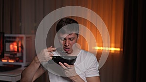 Handsome hungry young man eating spicy soup and trying to cool his mouth with hand sitting alone at table in evening or