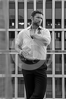 Handsome hunky man with unbuttoned shirt and loose bowtie stands on hotel balcony with sckyscraper backdrop photo