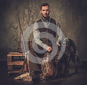 Handsome hunter with a english setter and shotgun in a traditional shooting clothing, sitting on a dark background.