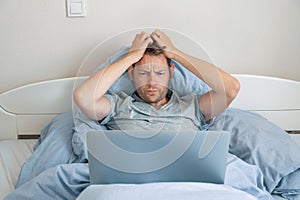 Handsome impressed man reading news on laptop with upset expression in bed in the morning. Guy amazed with bad news