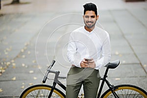 Handsome indian businessman in classic suit is using a smart phone and smiling while riding bicycle in city