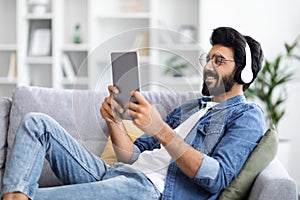 Handsome Indian Man In Wireless Headphones Using Digital Tablet At Home