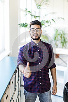 Handsome indian man working doing ok sign, thumb up with fingers, excellent sign standing in modern office