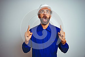 Handsome indian worker man wearing uniform and helmet over isolated white background amazed and surprised looking up and pointing