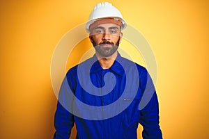 Handsome indian worker man wearing uniform and helmet over isolated yellow background with serious expression on face