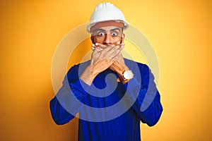 Handsome indian worker man wearing uniform and helmet over isolated yellow background shocked covering mouth with hands for