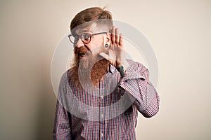 Handsome Irish redhead business man with beard wearing glasses over isolated background smiling with hand over ear listening an