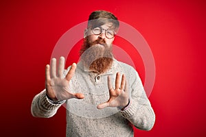 Handsome Irish redhead man with beard wearing casual sweater and glasses over red background Moving away hands palms showing