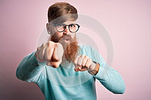 Handsome Irish redhead man with beard wearing glasses over pink  background Punching fist to fight, aggressive and angry