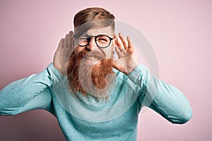 Handsome Irish redhead man with beard wearing glasses over pink isolated background Trying to hear both hands on ear gesture,