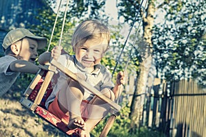 A handsome joyful boy of 2 years old is swinging on a swing and smiling. The older brother pushes the swing in which the younger b