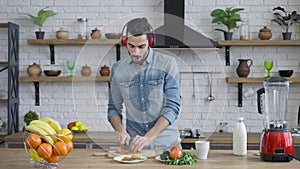Handsome joyful Middle Eastern man in headphone cutting tomato for healthful vegan salad and dancing. Portrait of