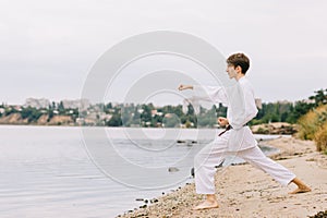 Beautiful boy in white karate kimono on the beach background. Judo practice concept. Copy space.