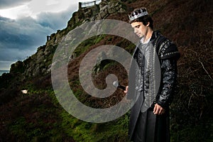 Handsome king with sword stands in contemplation with hill and parts of keep in the background