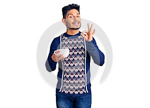 Handsome latin american young man drinking a cup of coffee doing ok sign with fingers, smiling friendly gesturing excellent symbol