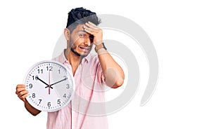 Handsome latin american young man holding big clock stressed and frustrated with hand on head, surprised and angry face