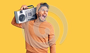 Handsome latin american young man holding boombox, listening to music angry and mad screaming frustrated and furious, shouting