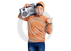 Handsome latin american young man holding boombox, listening to music surprised pointing with hand finger to the side, open mouth