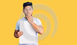 Handsome latin american young man holding cardboard banner with blank space covering mouth with hand, shocked and afraid for