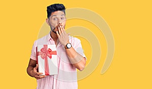 Handsome latin american young man holding gift covering mouth with hand, shocked and afraid for mistake