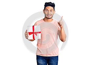 Handsome latin american young man holding gift surprised with an idea or question pointing finger with happy face, number one