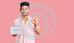 Handsome latin american young man holding heart calendar doing ok sign with fingers, smiling friendly gesturing excellent symbol