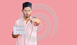 Handsome latin american young man holding heart calendar pointing with finger to the camera and to you, confident gesture looking