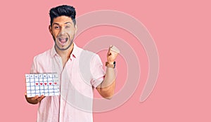 Handsome latin american young man holding heart calendar screaming proud, celebrating victory and success very excited with raised