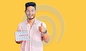 Handsome latin american young man holding heart calendar smiling happy and positive, thumb up doing excellent and approval sign