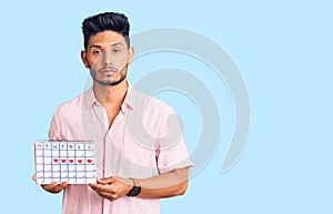 Handsome latin american young man holding heart calendar thinking attitude and sober expression looking self confident