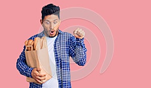 Handsome latin american young man holding paper bag with bread pointing down with fingers showing advertisement, surprised face