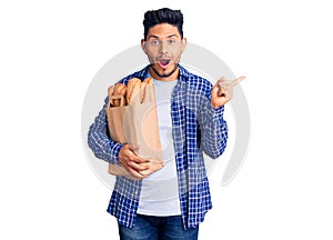 Handsome latin american young man holding paper bag with bread surprised pointing with finger to the side, open mouth amazed