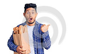 Handsome latin american young man holding paper bag with bread surprised pointing with hand finger to the side, open mouth amazed