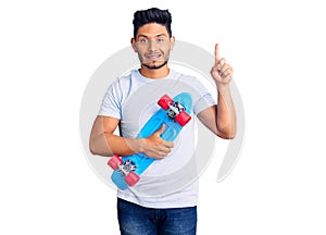 Handsome latin american young man holding skate surprised with an idea or question pointing finger with happy face, number one