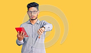 Handsome latin american young man holding touchpad with angry face, negative sign showing dislike with thumbs down, rejection