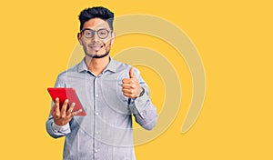 Handsome latin american young man holding touchpad smiling happy and positive, thumb up doing excellent and approval sign