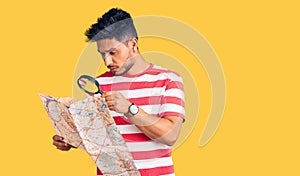 Handsome latin american young man looking at city map with magnifying glass thinking attitude and sober expression looking self