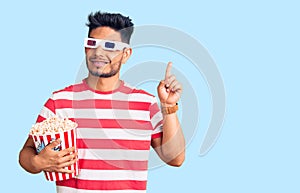 Handsome latin american young man wearing 3d glasses and eating popcorn at the movies surprised with an idea or question pointing