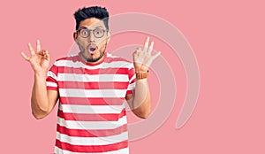 Handsome latin american young man wearing casual clothes and glasses looking surprised and shocked doing ok approval symbol with