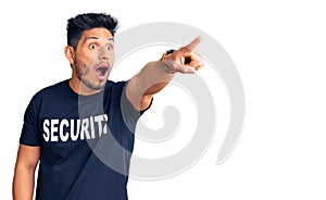 Handsome latin american young man wearing security t shirt pointing with finger surprised ahead, open mouth amazed expression,