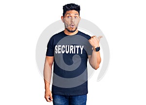 Handsome latin american young man wearing security t shirt surprised pointing with hand finger to the side, open mouth amazed