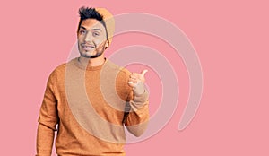 Handsome latin american young man wearing winter sweater and wool hat smiling with happy face looking and pointing to the side