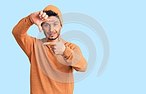 Handsome latin american young man wearing winter sweater and wool hat smiling making frame with hands and fingers with happy face