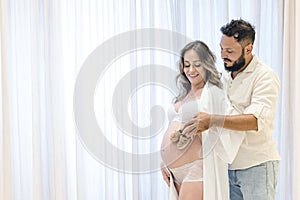 Handsome Latin man and his beautiful pregnant wife, couple holding children`s shoe over belly. Curtain background. pregnancy conce