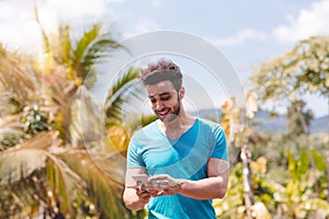 Handsome Latin Man Messaging With Cell Smart Phone Over Tropical Forest Background, Portrait Of Young Guy Chatting