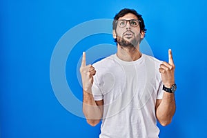 Handsome latin man standing over blue background amazed and surprised looking up and pointing with fingers and raised arms