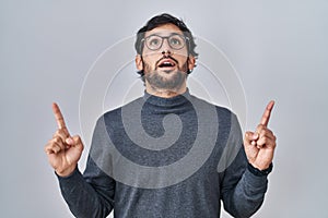 Handsome latin man standing over isolated background amazed and surprised looking up and pointing with fingers and raised arms