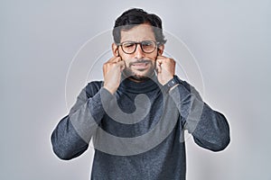 Handsome latin man standing over isolated background covering ears with fingers with annoyed expression for the noise of loud